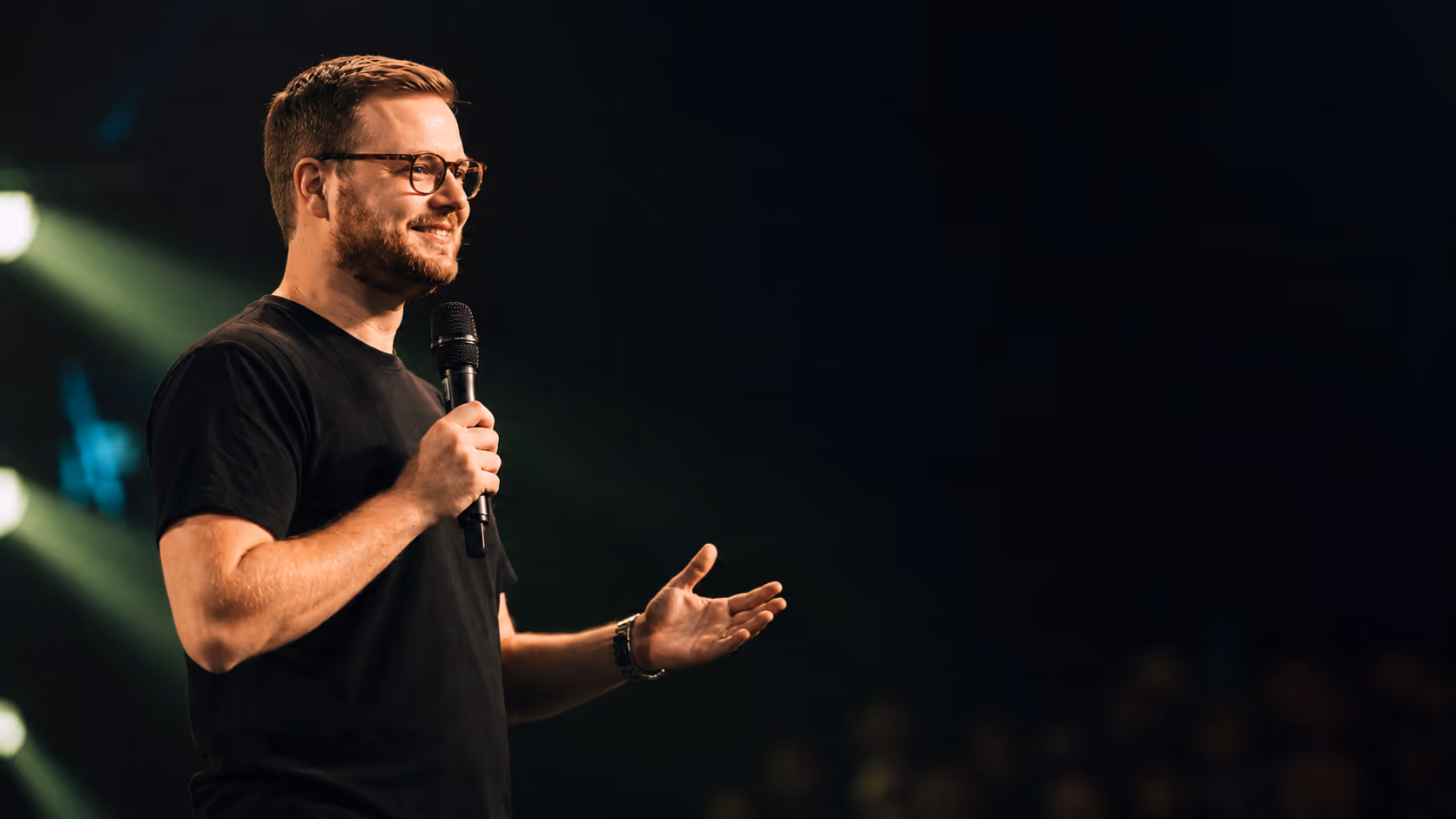 Man in glasses speaking into a microphone with hand gesturing, against dark backdrop.