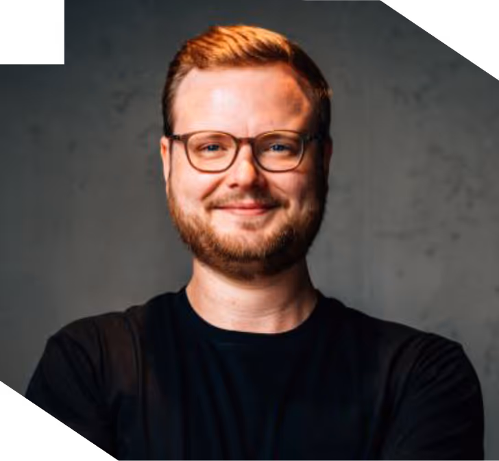 Smiling man with glasses, beard, and black shirt against a dark gray textured background.