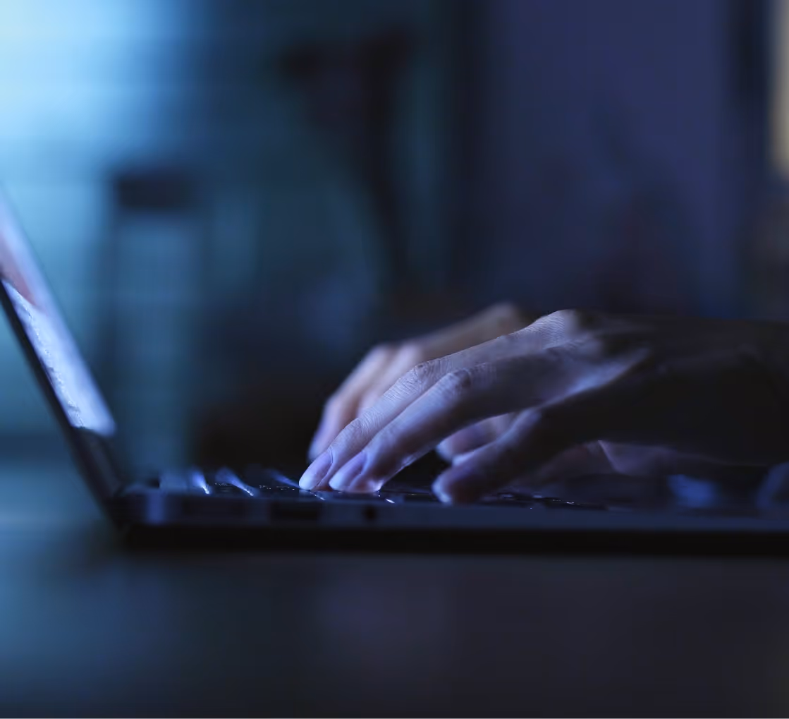 Hands typing on a laptop keyboard in a dimly lit room with blue ambient lighting.